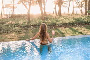 Una dona amb biquini al costat d'una piscina, amb una beguda, rodejada d'arbres i una cadira sàlva al fons.