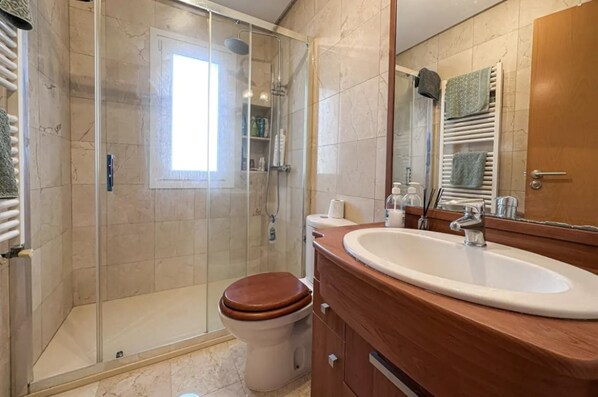 A bathroom with a glass-enclosed shower, a wooden toilet, a white sink, and a mirror.