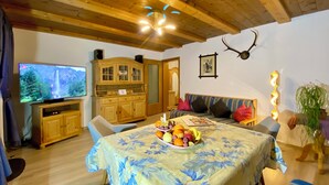 Een gezellige woonkamer met een houten plafond, een televisie, een eettafel met fruit, een bank en een aan de muur bevestigd hert.