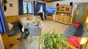 Een knus vertrek met een houten plafond, een eettafel met een tafelkleed met bloemenprint, een bank met gestreepte kussens, een televisie en een kast.