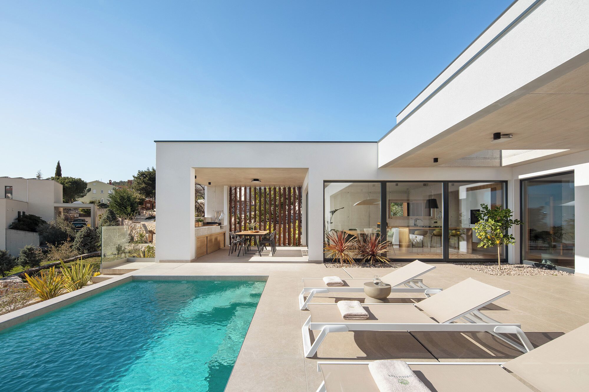 Une maison moderne dotée d’une piscine, d’un espace salon extérieur et d’une cuisine visible à travers des portes vitrées.