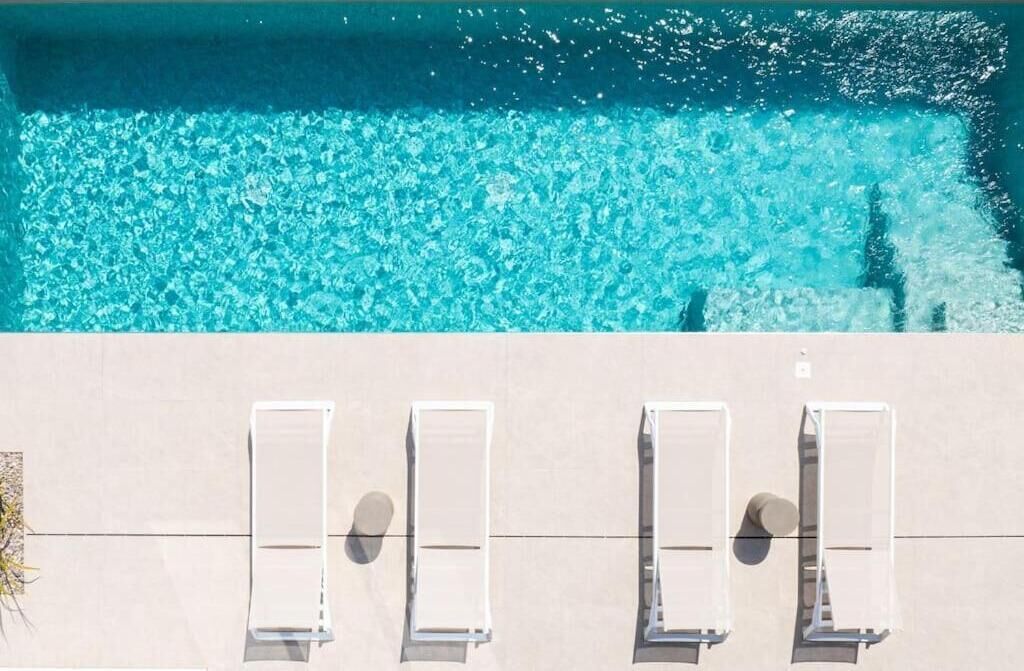 An aerial view of a swimming pool with clear blue water and four sun loungers aligned along the edge.