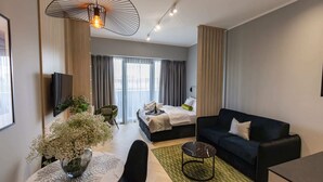 A modern hotel room with a bed, a sofa, a dining table with a vase of flowers, and a TV.