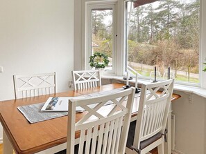 A dining area with a wooden table and white chairs, a window with a view of trees and a house, and a small plant on the windowsill. - 4 Star Holiday Home in Svanesund (Svanesund)