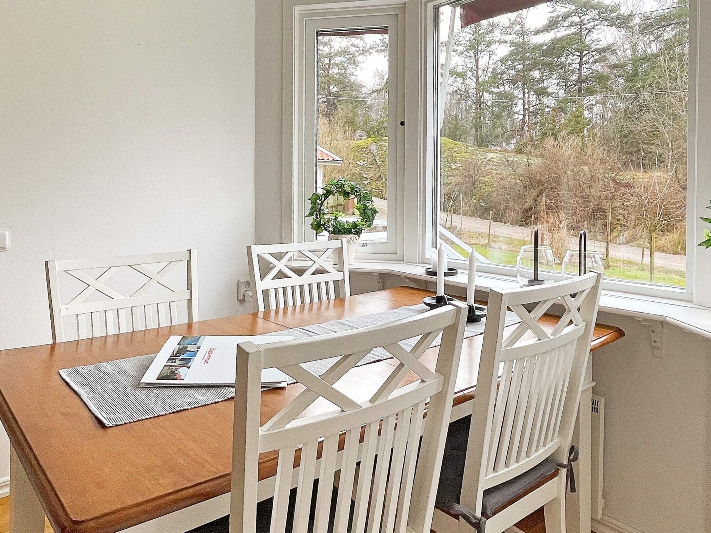 A dining area with a wooden table and white chairs, a window with a view of trees and a house, and a small potted plant on the windowsill.