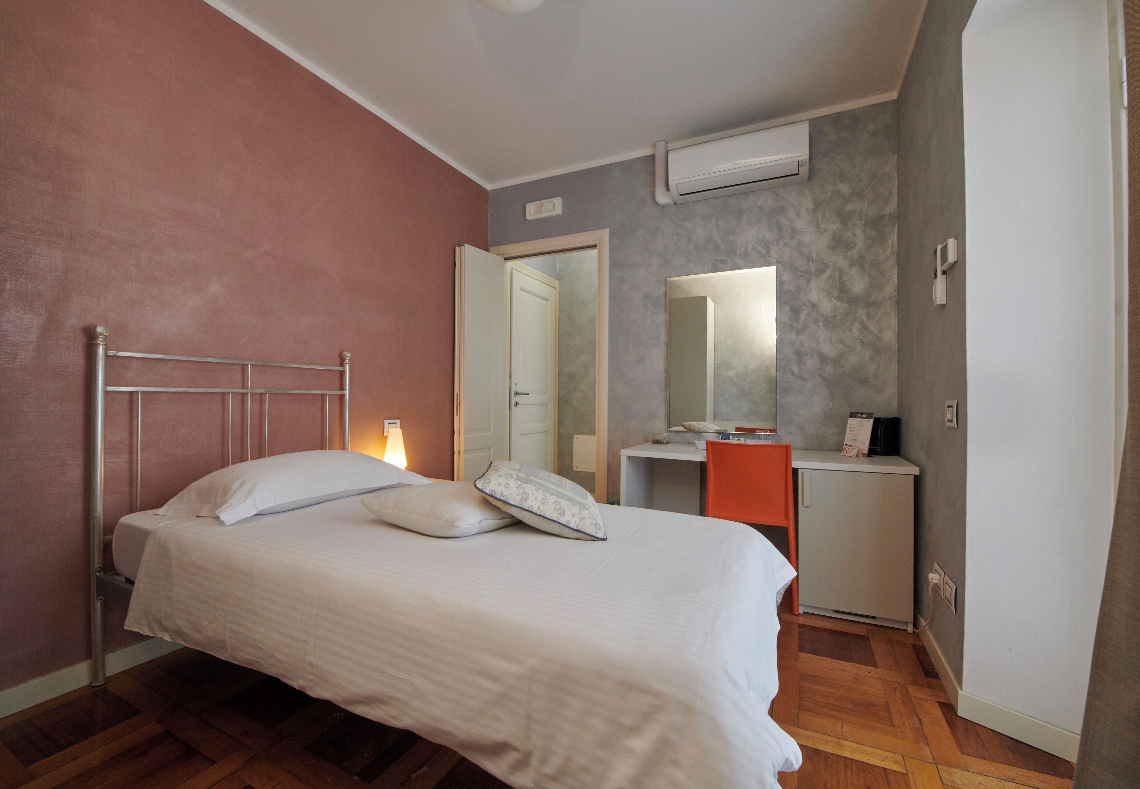 Ein Schlafzimmer mit einem Bett, einem Schreibtisch und einem Stuhl. Über dem Schreibtisch hängt ein Spiegel und an der Wand befindet sich eine Klimaanlage.