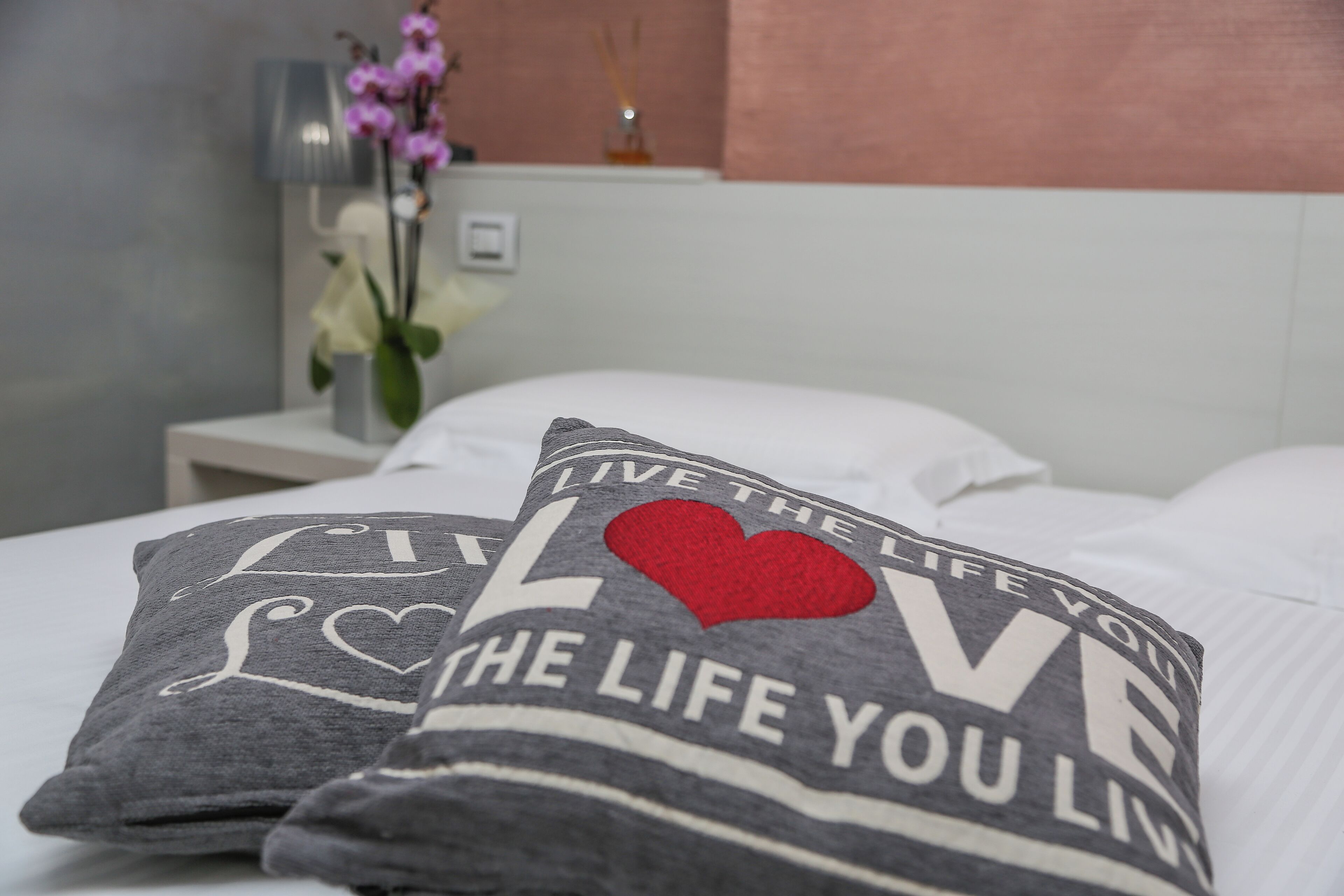 A bed with pillows that read "LIVE THE LIFE YOU LOVE" and a decorative vase with flowers in the background.