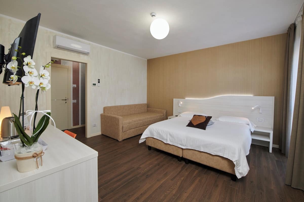 A hotel room with a bed, a sofa, a desk with a lamp, and an air conditioning unit.