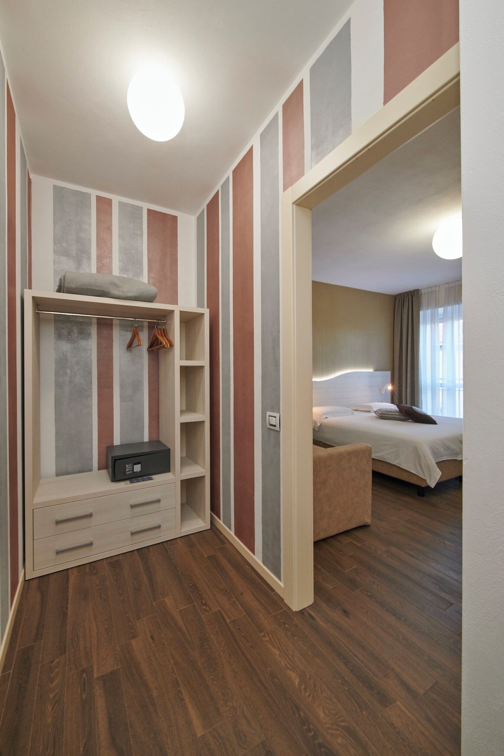 Habitación de hotel con piso de madera, cama con ropa blanca, mesita de noche, armario con espacio para colgar y un pequeño escritorio con cajones.
