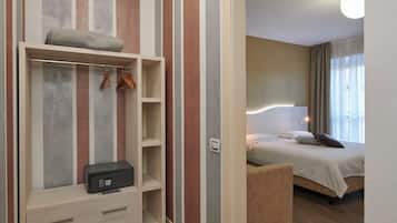 A hotel room with a wooden floor, a bed with white bedding, a bedside table, a wardrobe with hanging space, and a small desk with drawers.