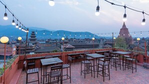 Un área de estar al aire libre con mesas y sillas, luces colgantes y vistas a un paisaje urbano con montañas al fondo.
