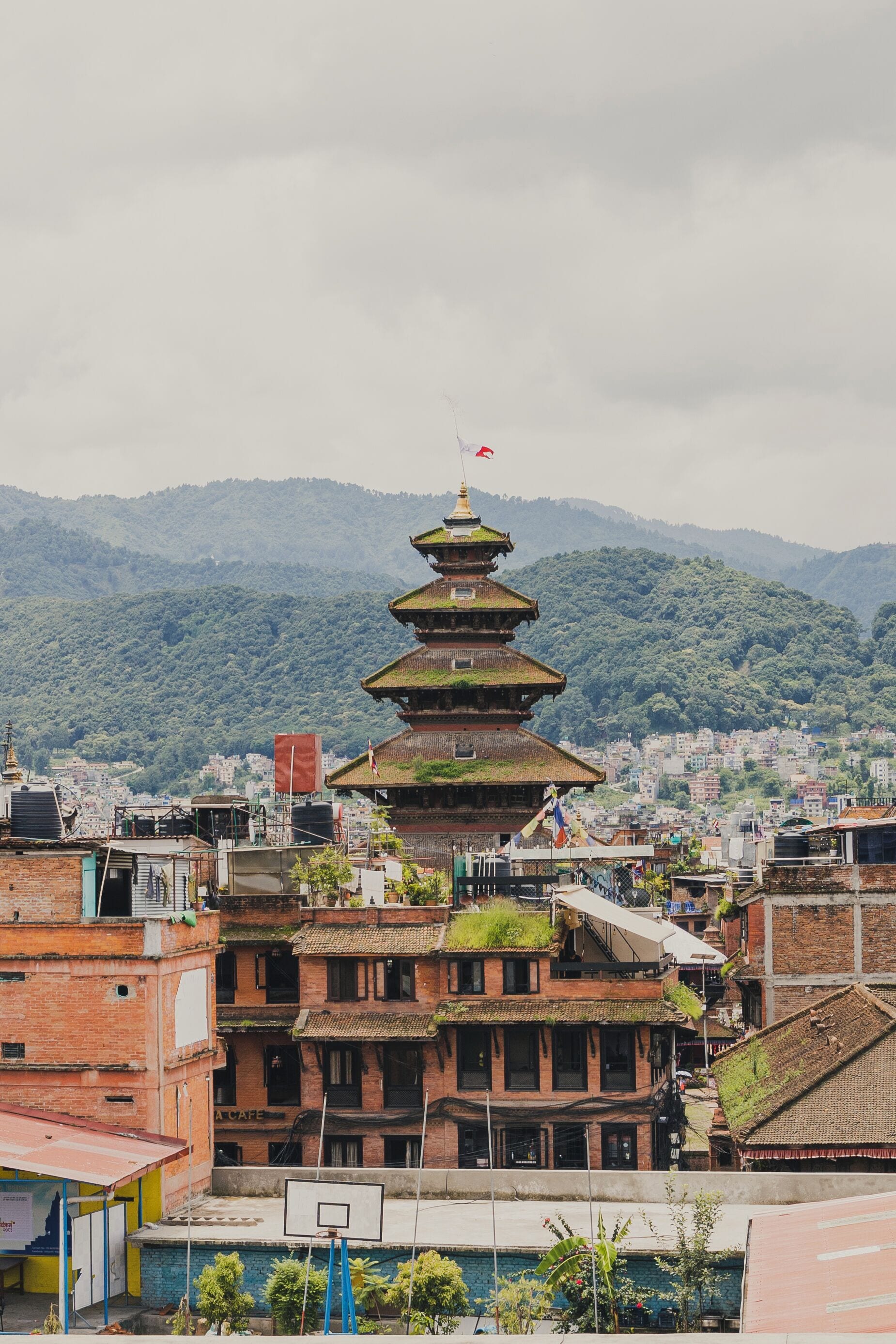 Una pagoda tradicional de varios niveles, con una bandera en la parte superior, rodeada de edificios urbanos y un paisaje montañoso.