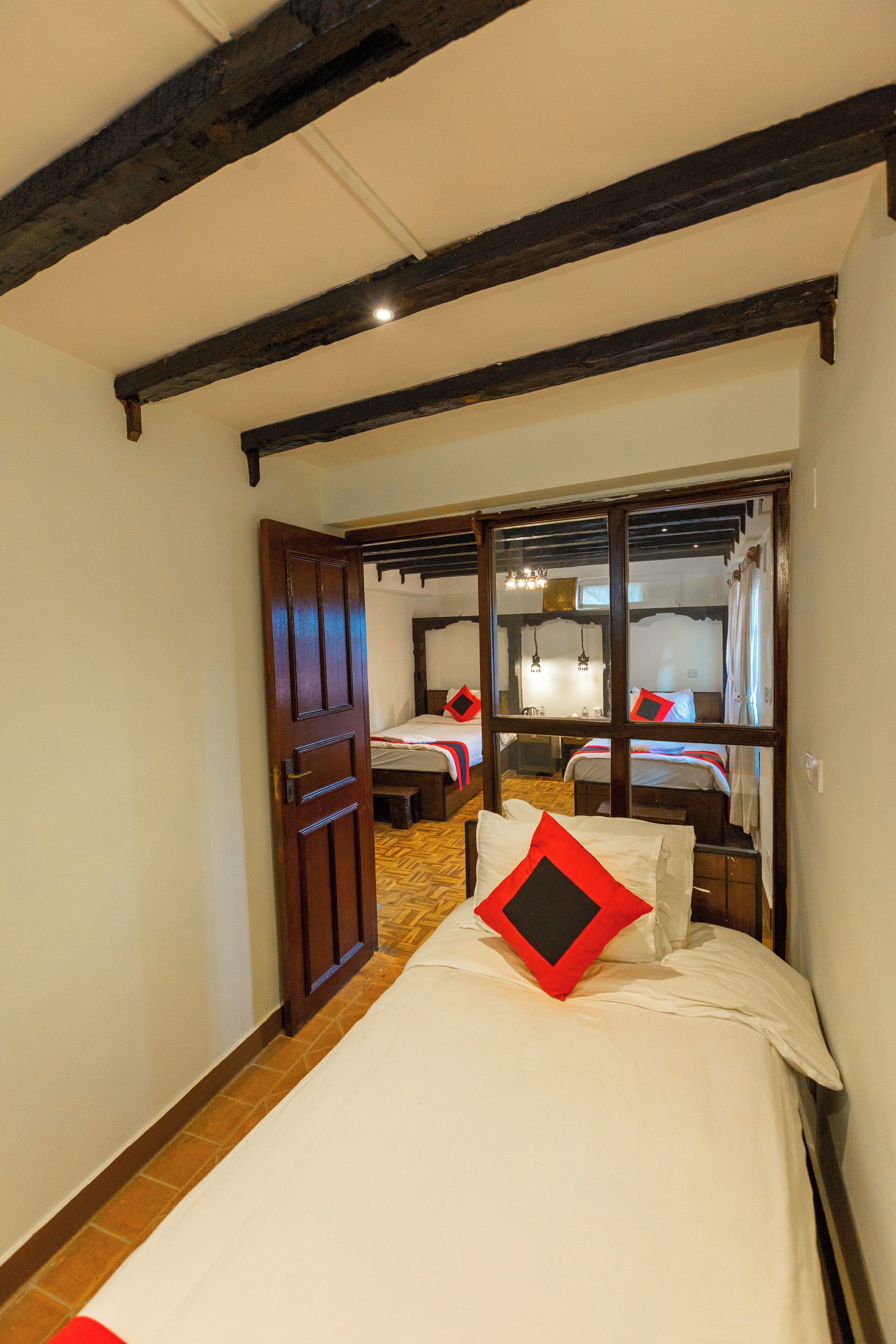 A room with a wooden door, a bed with white bedding and a red pillow, and a mirror reflecting another room with a bed and a hanging light fixture.
