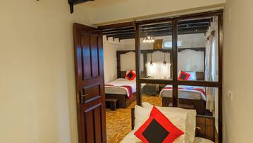 A room with a wooden door, a bed with white bedding and a red pillow, and a mirror reflecting another room with a bed and a hanging light fixture.
