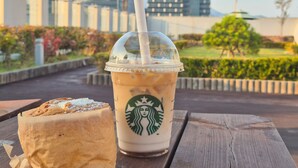 Un vaso de Starbucks con una pajita sobre una mesa de madera, un pastel y un paisaje urbano al fondo.