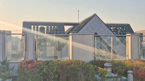 Un edificio moderno con fachada de vidrio, techo metálico y un jardín con arbustos y una fuente decorativa.