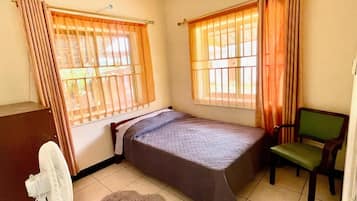 A small room with a bed, a chair, a desk, and a fan.