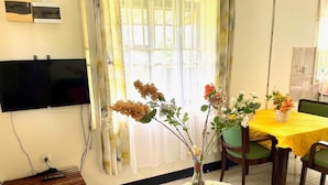 A room with a TV mounted on the wall, a wooden bench, a round table with a yellow tablecloth, and a vase with flowers. - Tropical Paradise in Paramaribo 3 - Room House (Paramaribo)