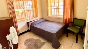 A small, single-bed room with a bed, a chair, a fan, and a window with curtains. - Tropical Paradise in Paramaribo 3 - Room House (Paramaribo)