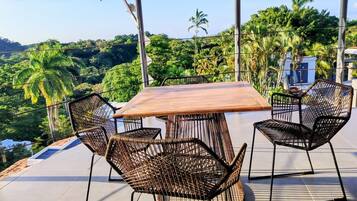 An outdoor patio with a wooden table and wicker chairs, surrounded by lush greenery and a clear view of the landscape.