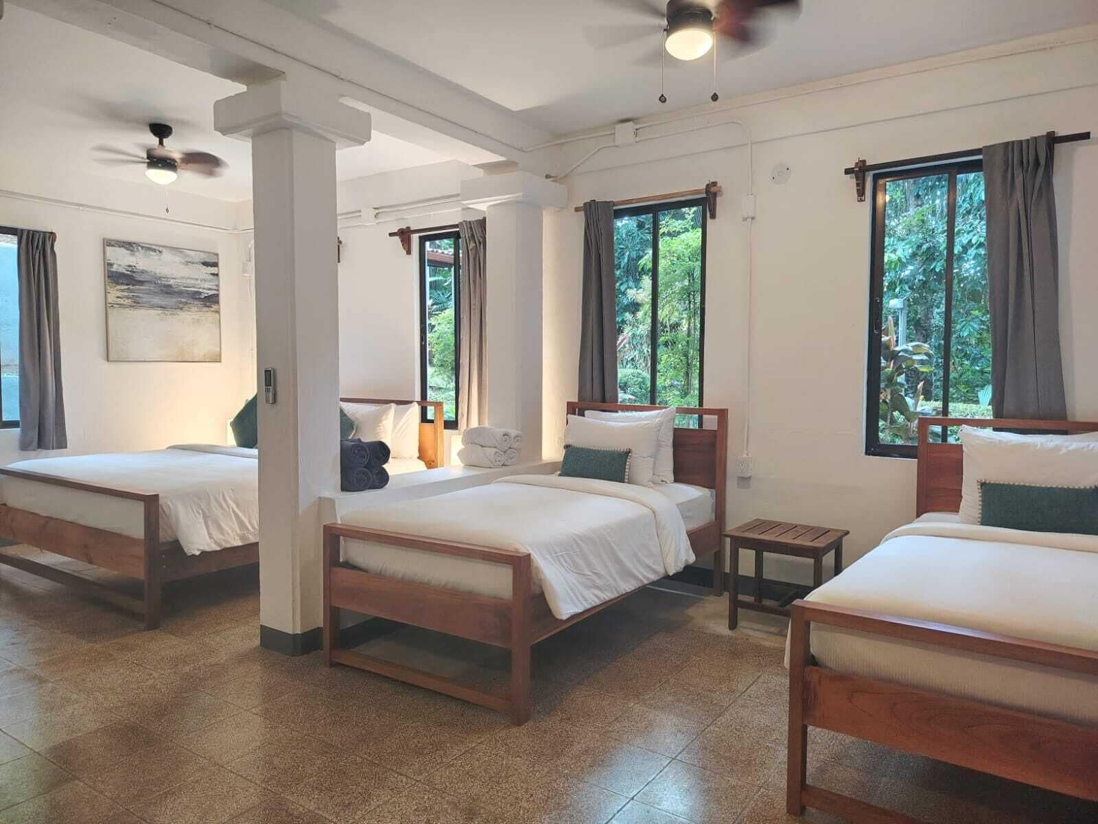 Una camera d'albergo con tre letti in legno, un ventilatore a soffitto e finestre con tende.