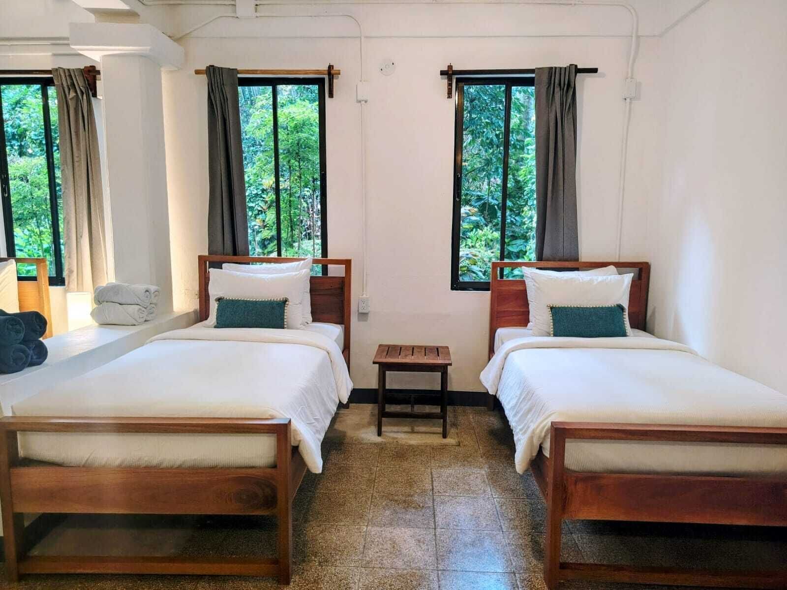 Una habitación con dos camas de madera, una mesita y ventanas con cortinas.