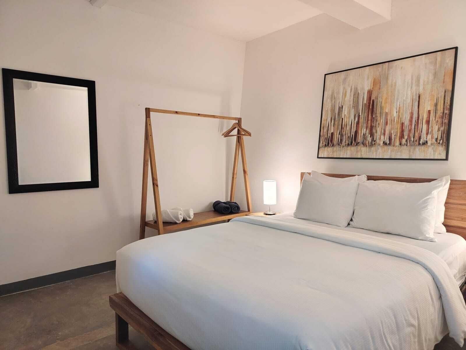 A bedroom with a bed, a wooden swing, a bedside table with a lamp, and a large abstract painting on the wall.