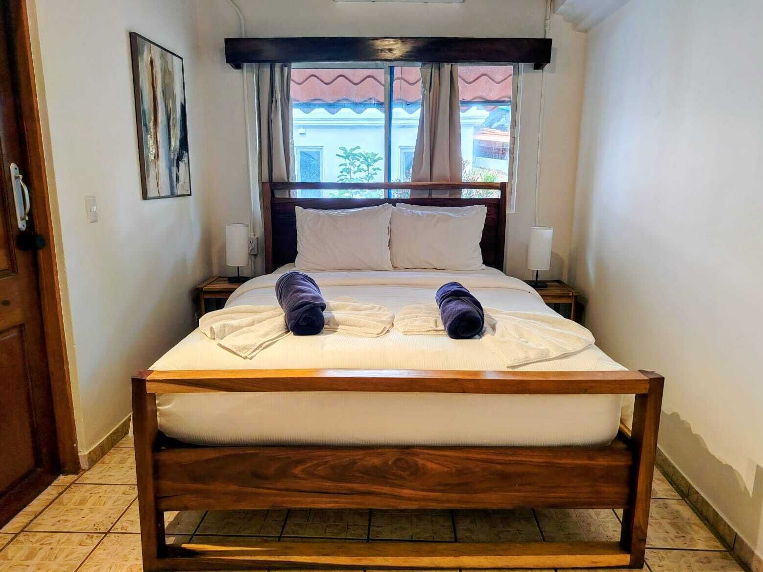 A hotel room with a wooden bed, white linens, two rolled towels, a nightstand with a lamp, and a window with curtains.