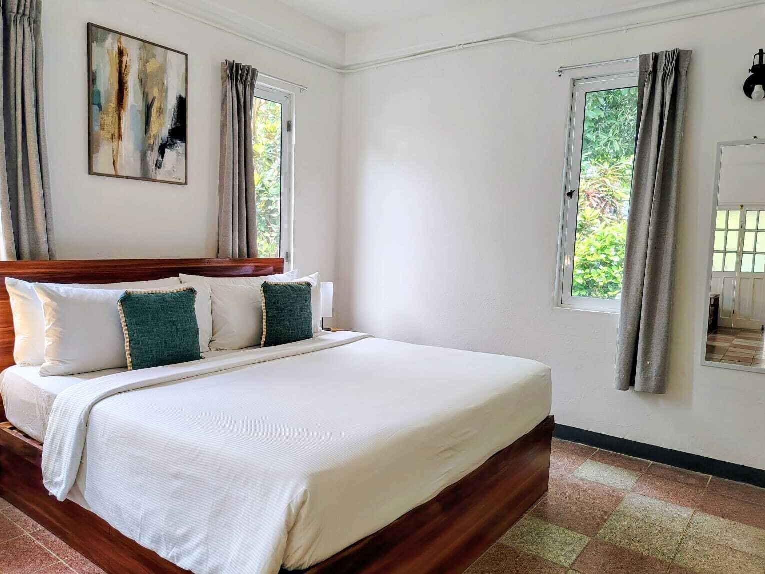 A neatly made bed with a wooden headboard, two windows with curtains, and a painting on the wall.