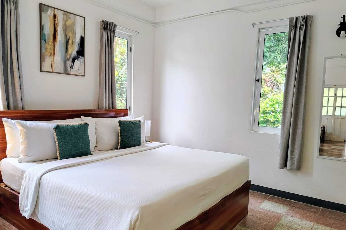A neatly made bed with a wooden headboard, two windows with curtains, and a painting on the wall.