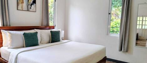 A bedroom with a large bed, two windows with curtains, a painting on the wall, and a view of greenery outside.