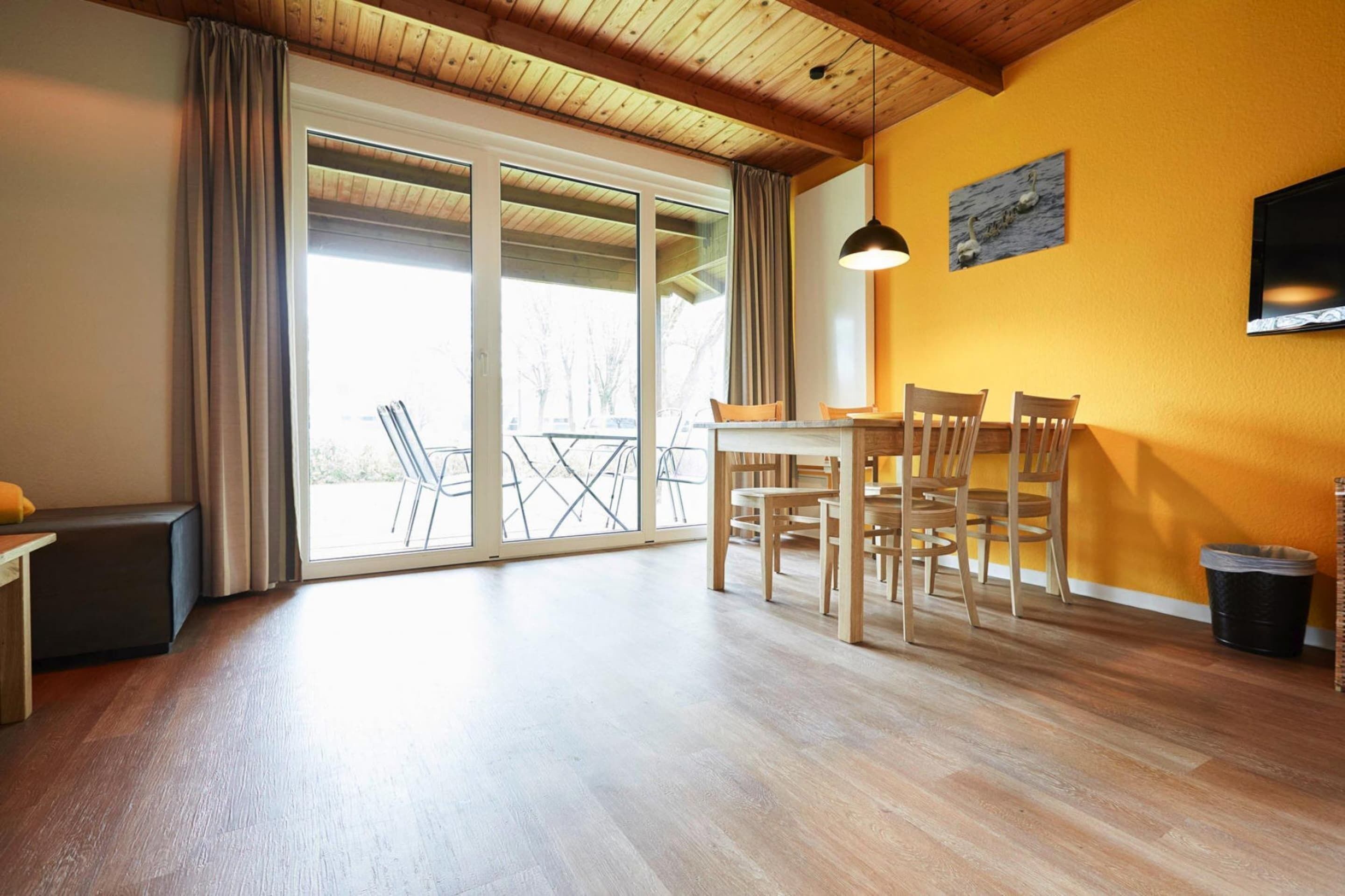 A spacious room with a wooden floor, a dining table with chairs, a bench, and a large sliding glass door leading to an outdoor area with a view of trees.