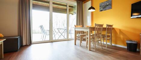 A spacious room with a wooden floor, a dining table with chairs, a bench, and a large sliding glass door leading to an outdoor area with a view of trees.