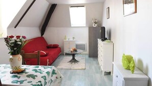A small, well-lit room with a sloped ceiling, a red sofa, a small table with a vase of flowers, a white dresser, and a window providing natural light.