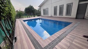 Una piscina rectangular con un borde de madera, una escalera metálica y una casa con ventanas amplias y una puerta al fondo.