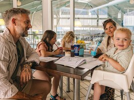 En familie spiser ved et bord med papirer, drikkevarer og udsigt til det fri.