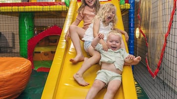 Children sliding down a yellow slide in a play area with colorful netting and a soft orange seating area.