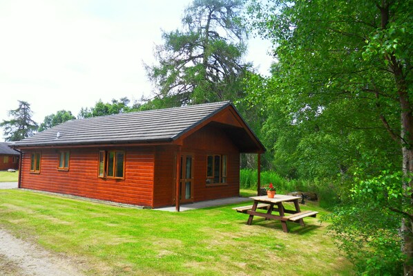 A wooden cabin with a picnic table in a grassy area surrounded by trees.