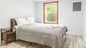 A bedroom with a bed, a nightstand, a window with blinds, and a wall-mounted electrical outlet. - Charming cottage Pulaski VIP Rentals 1.8 miles away from famous salmon river (Pulaski)