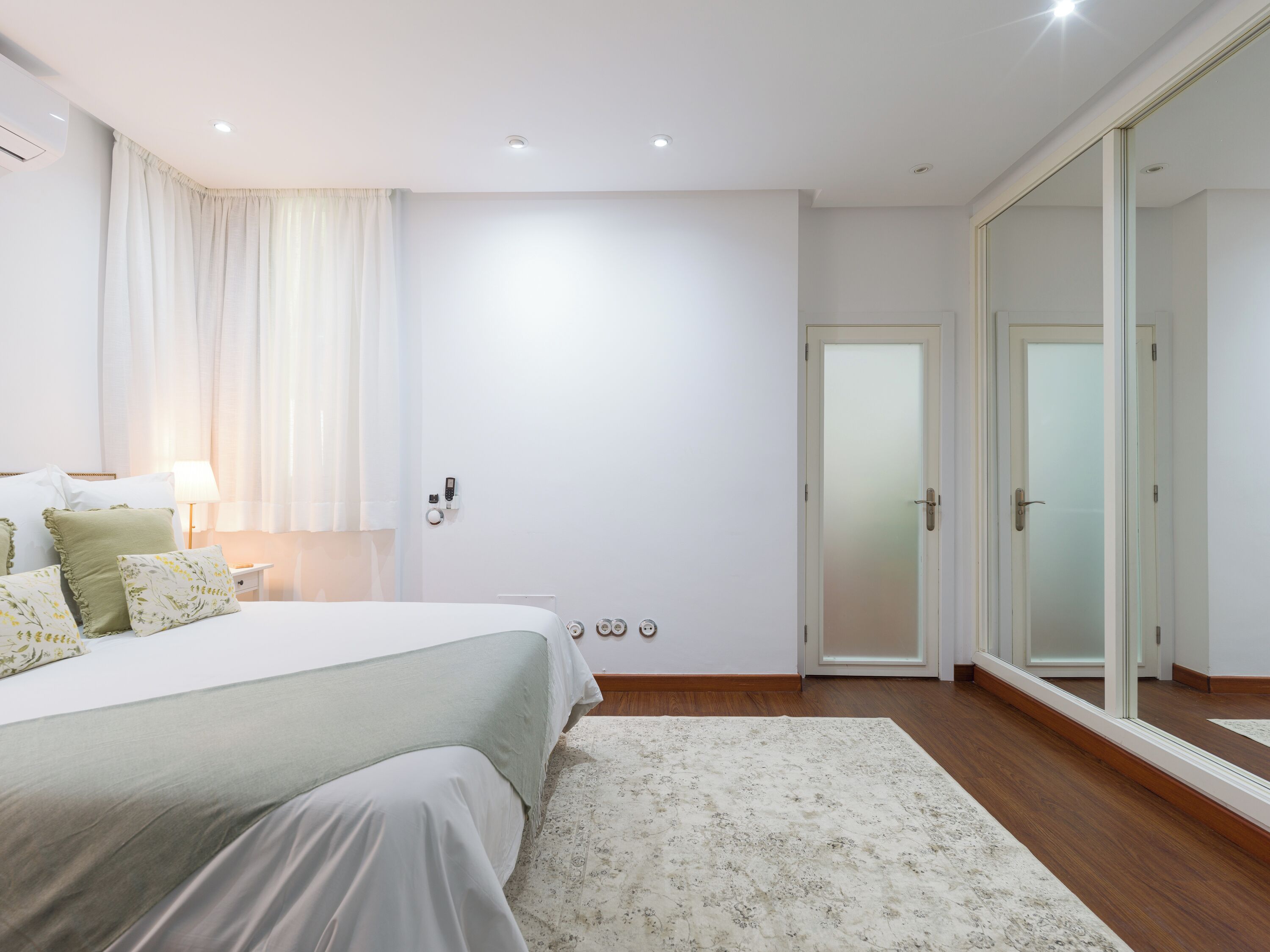 A modern bedroom with a large bed, a bedside table with a lamp, a wall-mounted phone, and a sliding glass door leading to a bathroom.