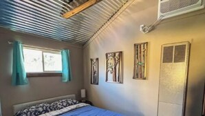 Ein Schlafzimmer mit einem Bett, einem Fenster mit Vorhängen, Wandbildern und einer Klimaanlage.