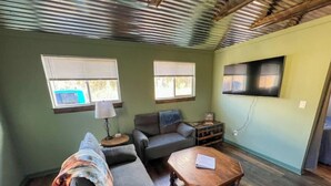 Ein gemütliches Wohnzimmer mit einer Couch, einem Sessel und einem Holztisch. Zwei Fenster mit Jalousien, eine Lampe und ein Flachbildfernseher an der Wand sind zu sehen.