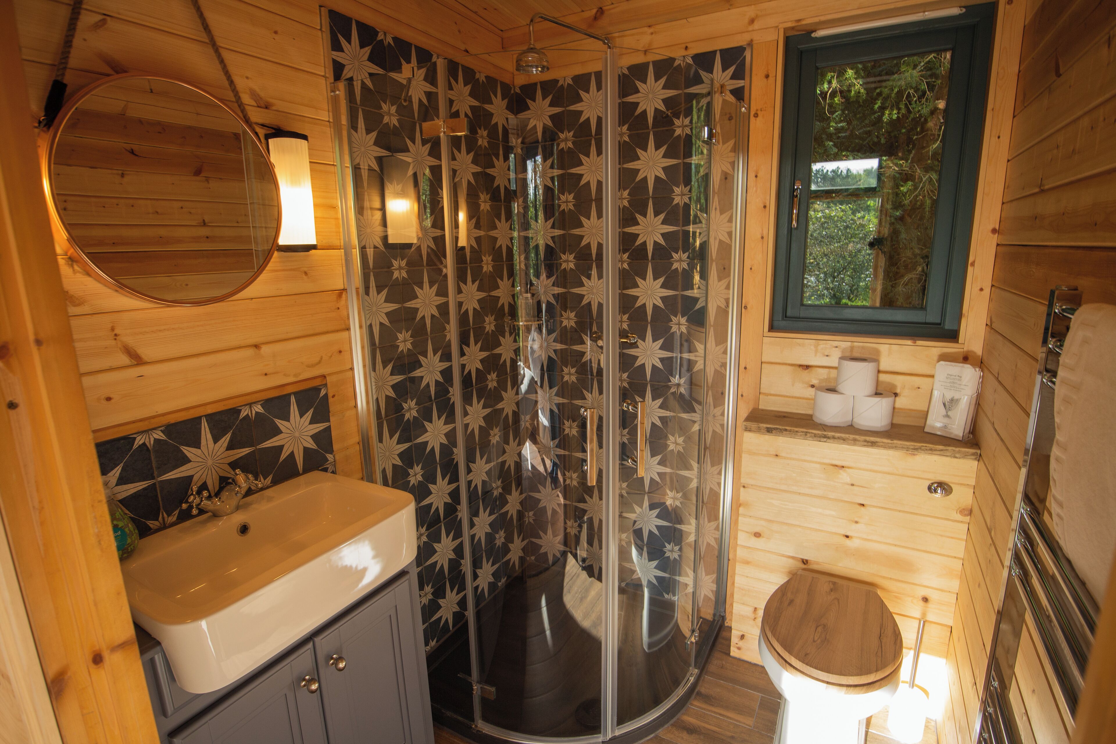 A wooden cabin bathroom with a shower, sink, toilet, and a window with a view of trees.