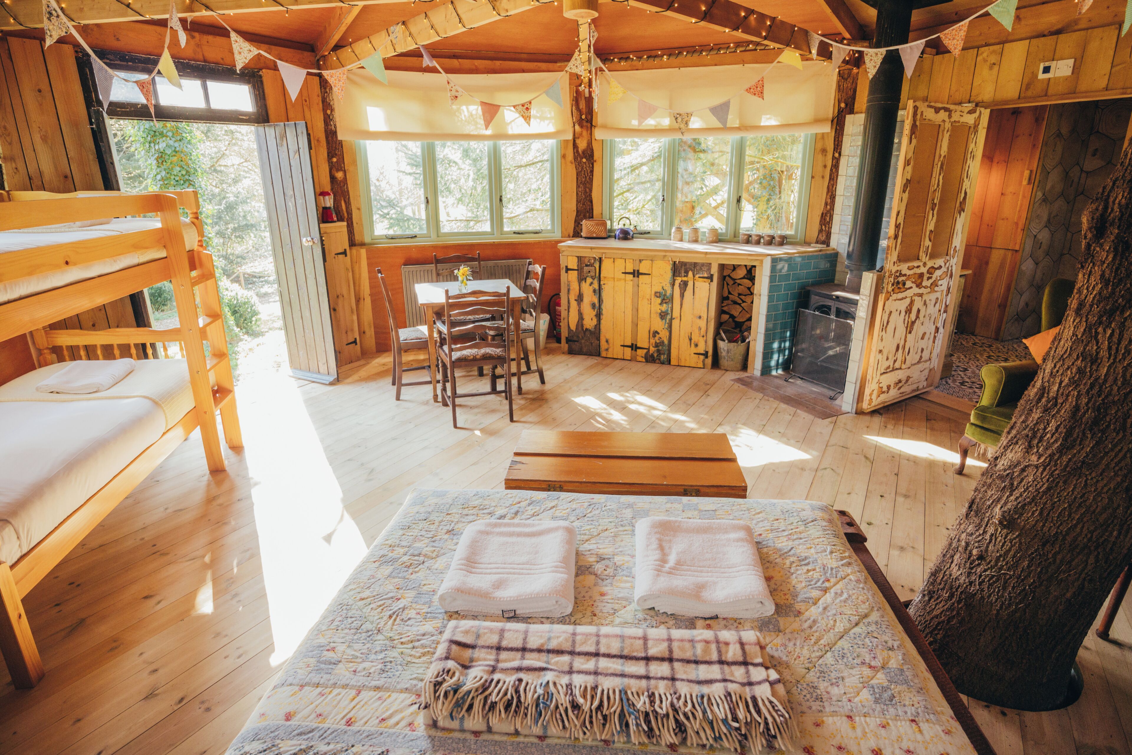Interior rústico de una cabaña con una cama litera de madera, una mesa de comedor con sillas, una zona de cocina y un gran tronco de árbol.