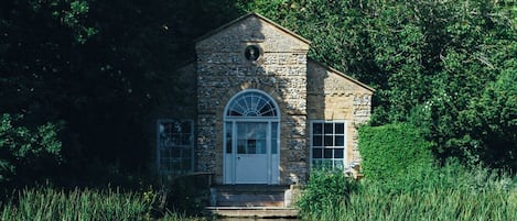 A stone building with a white door and windows, surrounded by lush greenery and water.