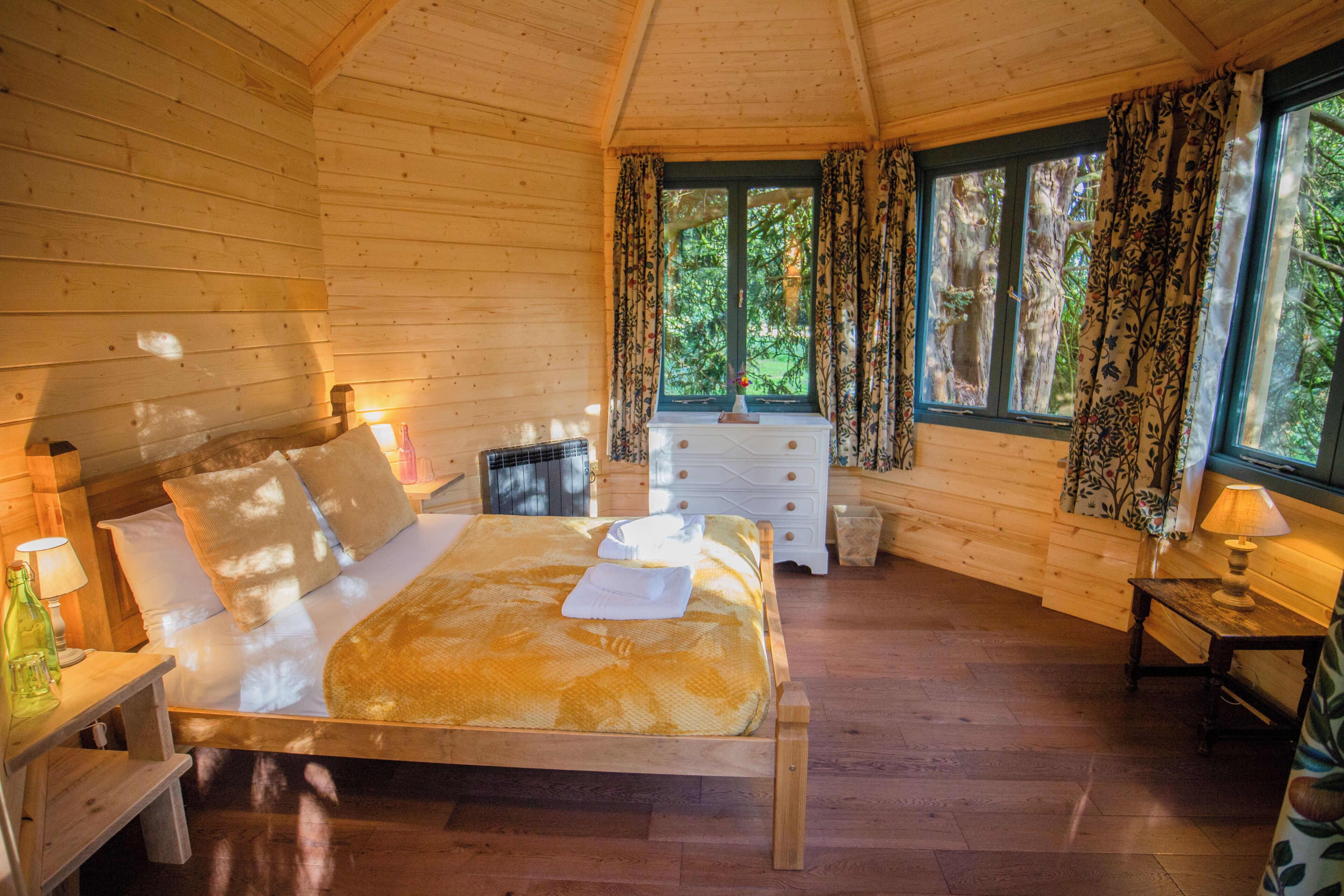 A wooden cabin bedroom with a bed, dresser, and window with curtains.