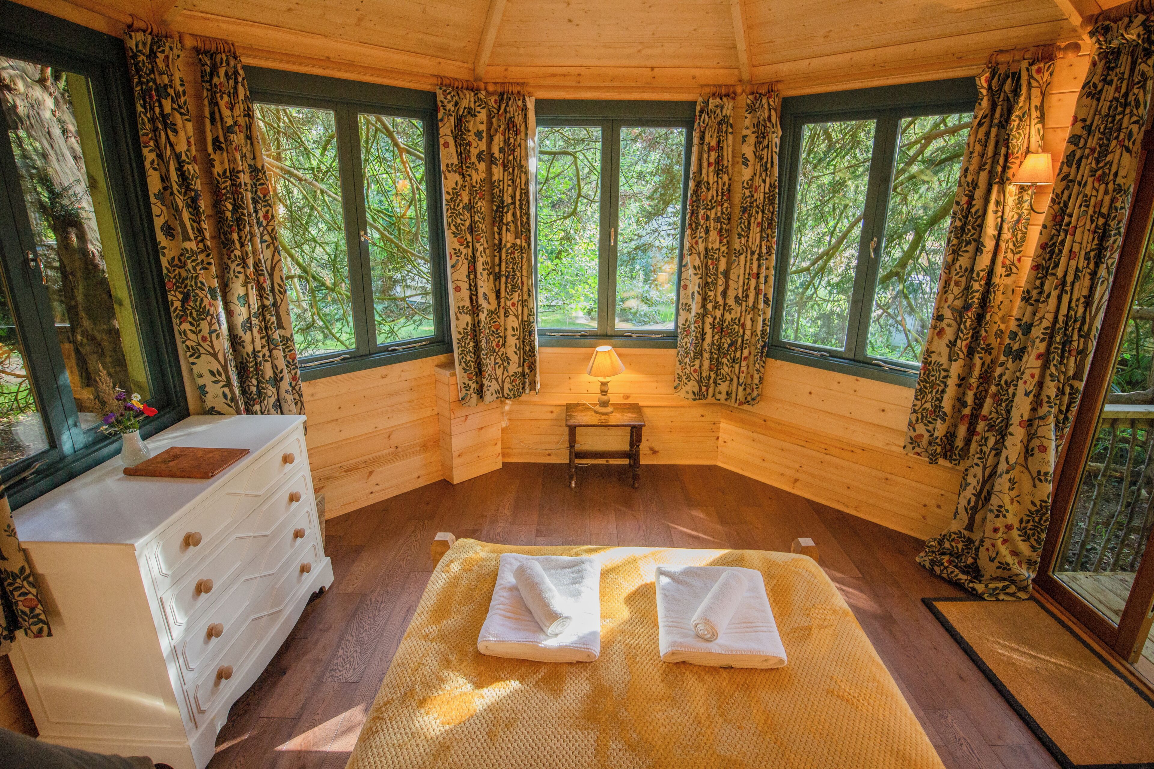 Una cabaña con paredes de madera, una cama con toallas, una lámpara y una ventana con cortinas.