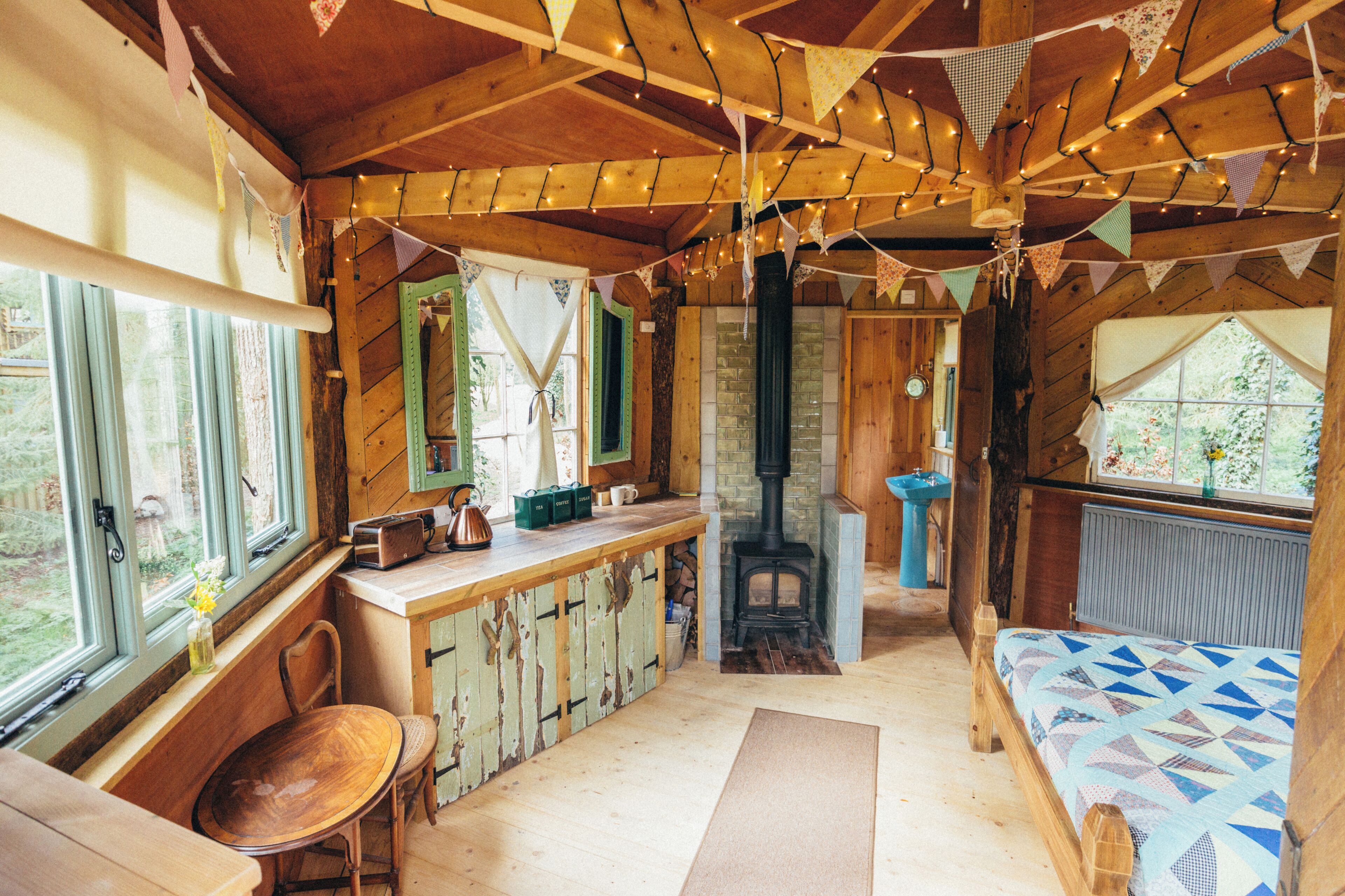 Interior rústico de una cabaña con comedor de madera, chimenea central, cama con colcha de estampado geométrico y ventana con vistas a la vegetación.