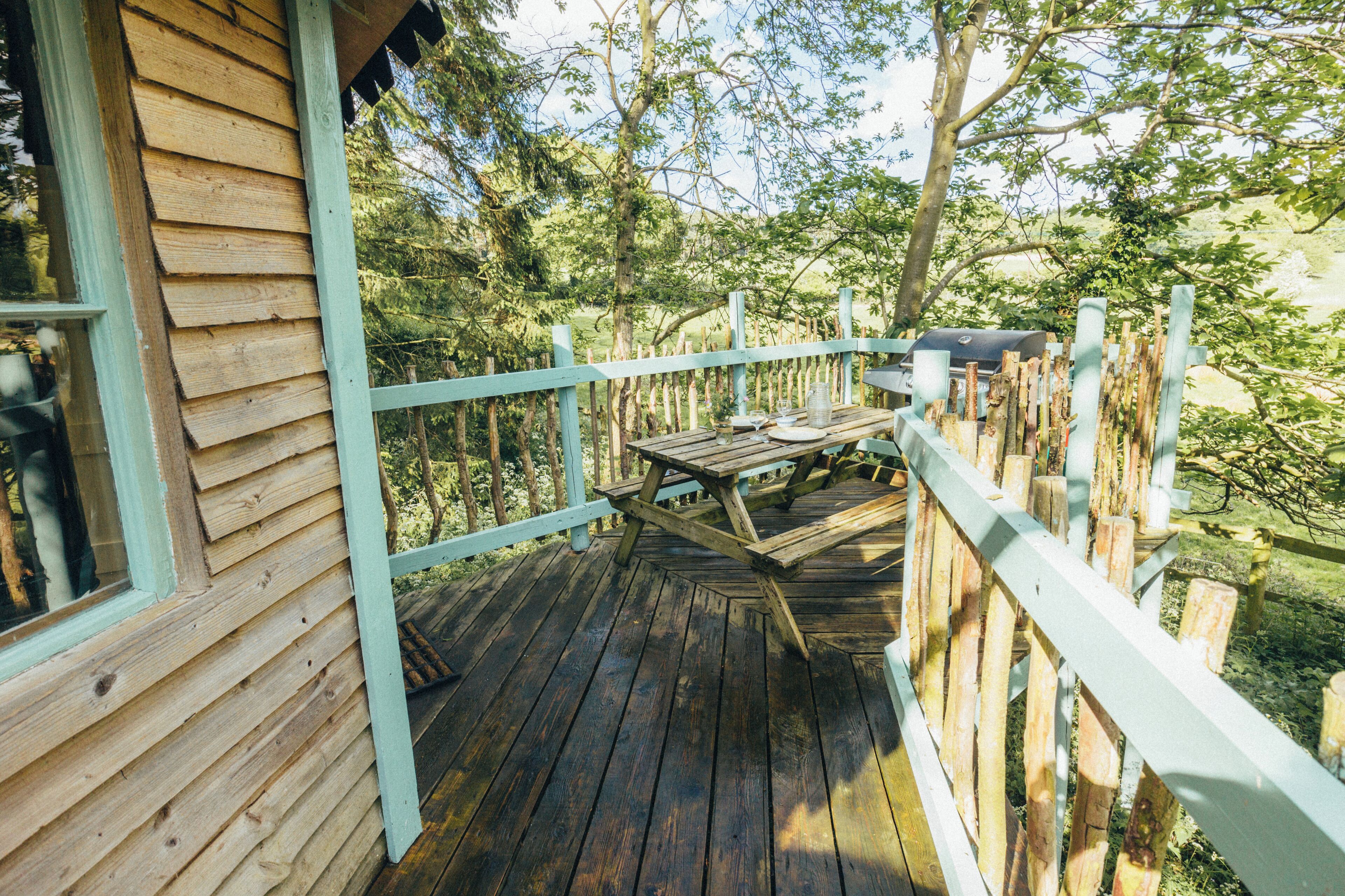 A wooden deck with a table and chairs, surrounded by a light blue railing, overlooking a forested area.