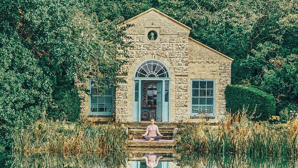 A stone building with a central arched window and a person meditating on the steps. - West Lexham Wellness and Retreat Centre (King's Lynn)